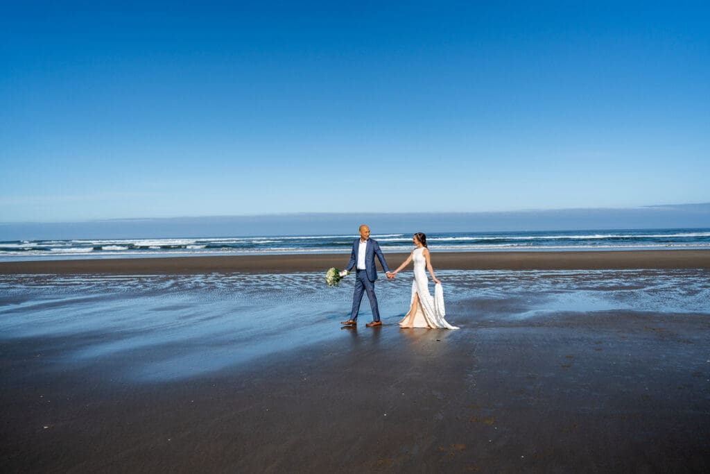 Couple walking hand in hand along Arch Cape beach with the ocean and cliffs in the background during their Oregon Coast elopement.